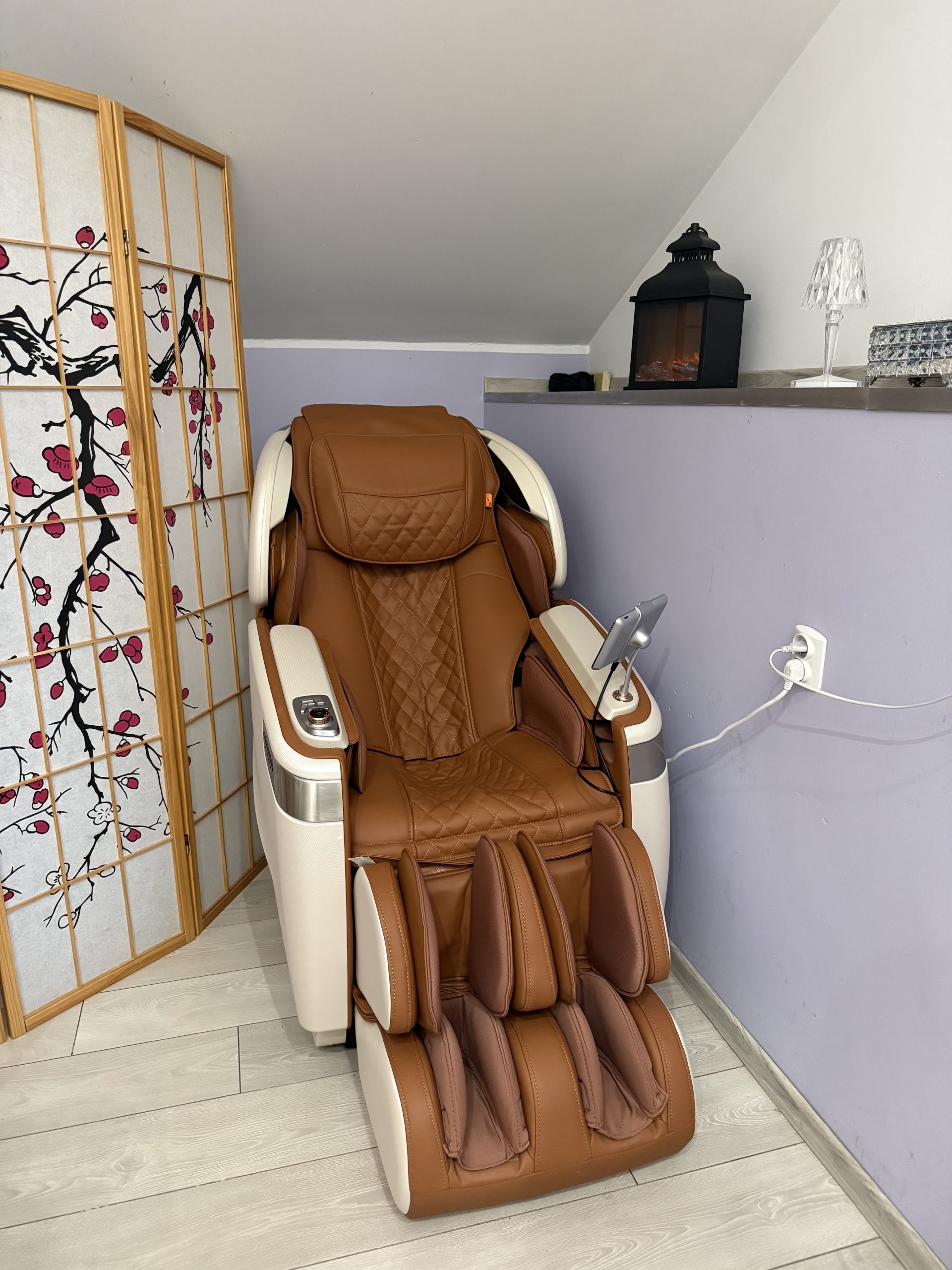 Brązowo-beżowy, luksusowy fotel masujący ze skóry stoi w rogu jasnego pomieszczenia gabinetu spa. Obok fotela znajduje się składany parawan z motywem kwitnącej wiśni i metalowa latarenka.