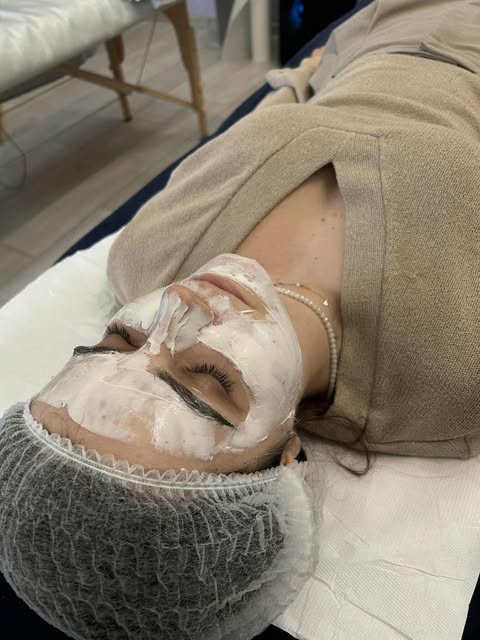 Kobieta leży na leżance zabiegowej w czepku jednorazowym i szlafroku, z białą, grubą maską kosmetyczną nałożoną na twarz. Jej oczy są zamknięte, a maska pokrywa całą twarz, omijając tylko usta.